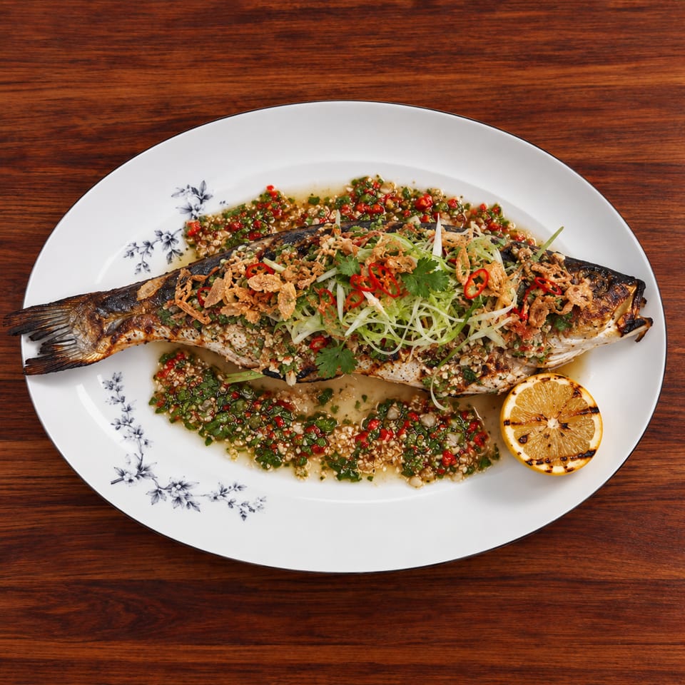 Whole grilled fish topped with herbs, sliced chilies, crispy shallots, and sauce, served with a grilled citrus half.