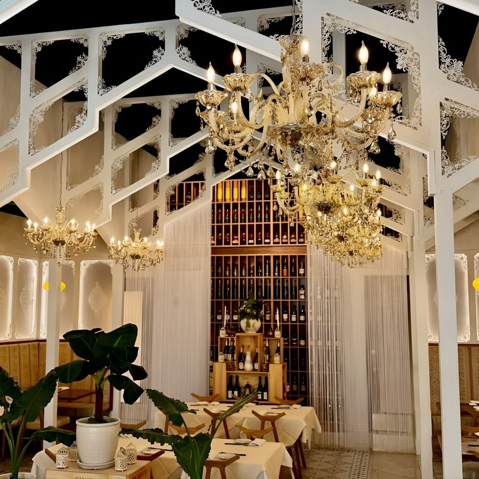 Elegant restaurant interior with ornate white ceiling, sparkling chandeliers, and a large wine bottle display.